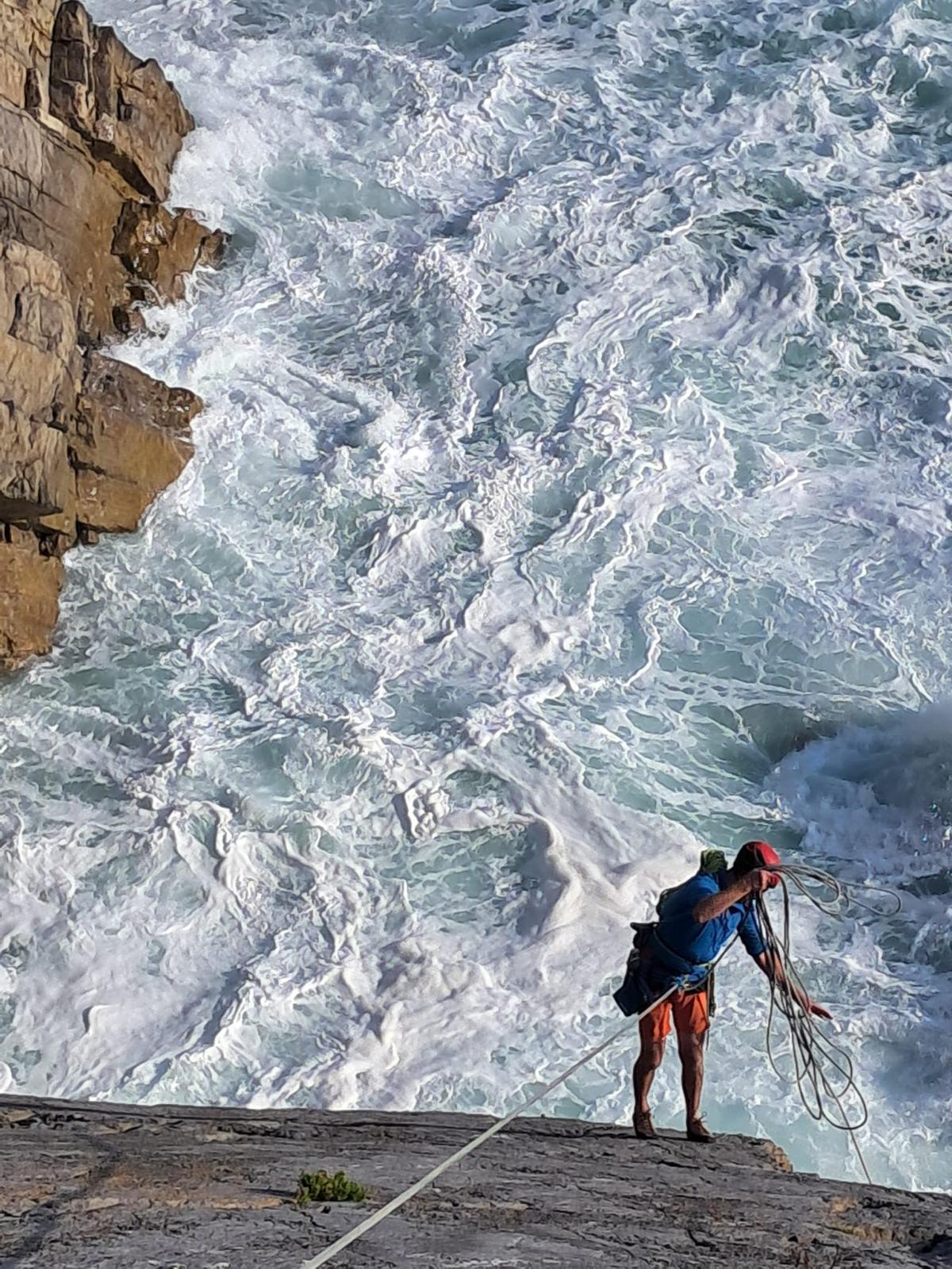 A club member in a red helmet flaking a rope on the clifftop, with surf breaking against the rock far below.
